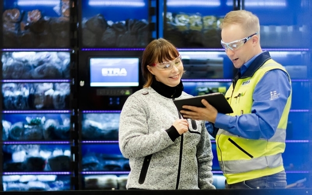 En Etra-medarbetare presenterar en automationsl&ouml;sning f&ouml;r en kund i en lagermilj&ouml;, i bakgrunden ett f&ouml;rdelningssk&aring;p m&auml;rkt med ETRA-logotypen.