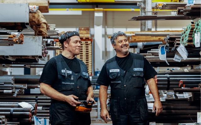 Two smiling factory workers wearing dark blue uniforms with name tags standing in a warehouse or metal workshop, surrounded by metal rods and storage racks.