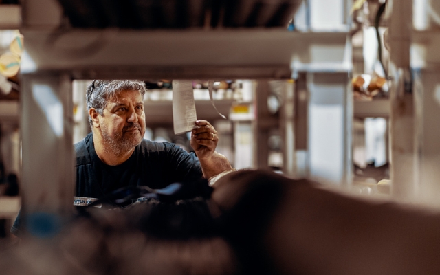 Man i arbetsuniform som inspekterar eller hämtar en etikett eller följesedel från en hylla i ett industriellt lager eller förvaringsutrymme, sedd genom hyllsektioner.