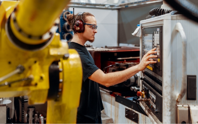 Fabrikarbetare med skyddsglasögon och hörselskydd som styr en CNC-maskin via en kontrollpanel, med en stor gul industrirobotarm i förgrunden.