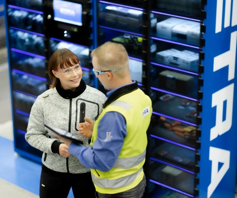 En Etra-medarbetare presenterar en automationsl&ouml;sning f&ouml;r en kund i en lagermilj&ouml;, i bakgrunden ett f&ouml;rdelningssk&aring;p m&auml;rkt med ETRA-logotypen.