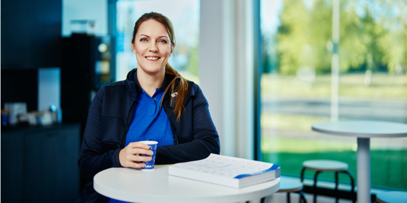 En leende kvinna i Etras bl&aring; profilkl&auml;der sitter vid ett litet bord med en kaffekopp och en broschyr.
