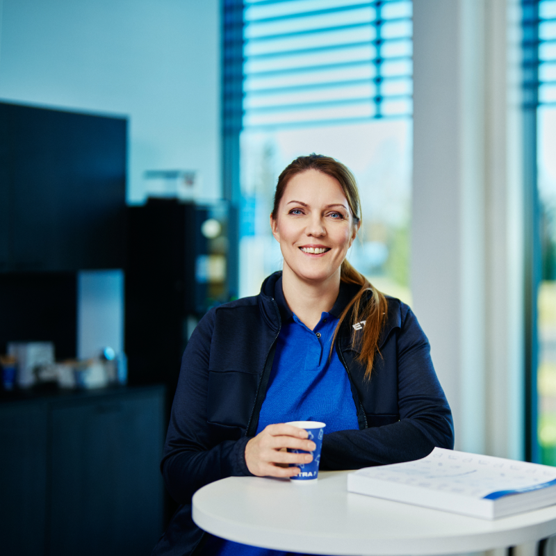 En leende kvinna i Etras bl&aring; profilkl&auml;der sitter vid ett litet bord med en kaffekopp och en broschyr.