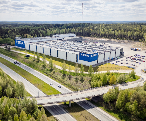 Flygbild av Etras logistikcenter, en stor vit- och bl&aring;randig industribyggnad med ETRA-logotyper p&aring; fasaden, omgiven av skog och bel&auml;gen intill en motorv&auml;g med en bro.