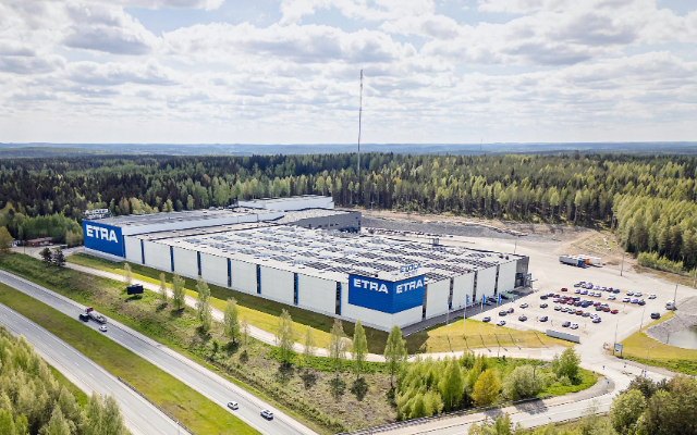 Flygbild av Etras logistikcenter, en stor vit- och bl&aring;randig industribyggnad med ETRA-logotyper p&aring; fasaden, omgiven av skog och bel&auml;gen intill en motorv&auml;g med en bro.