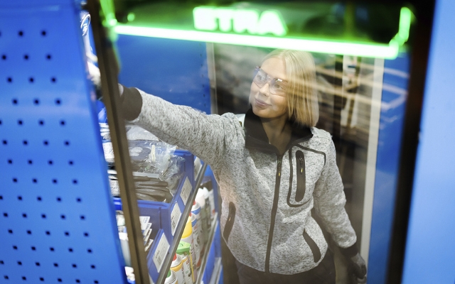 En kvinna i arbetskläder plockar en produkt från en hylla i ett lager eller serviceutrymme. Hon bär skyddsglasögon och ett grått fleeceplagg. Reflektionen visar en grön skylt med texten "ETRA".