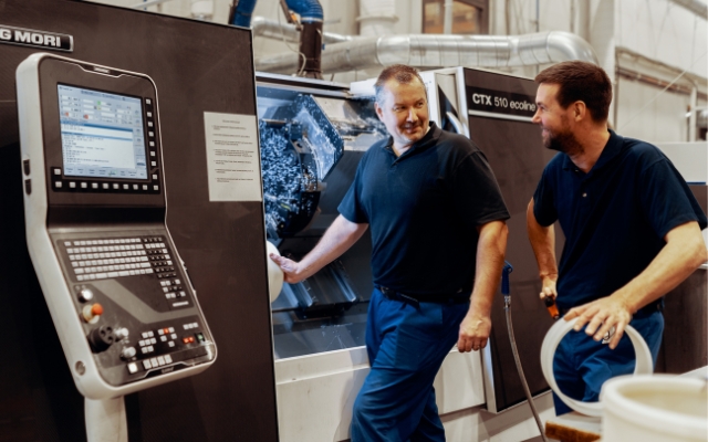 Två män i mörkblå arbetskläder arbetar vid en CNC-maskin i en industrimiljö. Den ena mannen står vid maskinens öppning medan den andra håller i ett rör. Till vänster syns ett styrpanel med skärm och knappar märkt 'DMG MORI'.