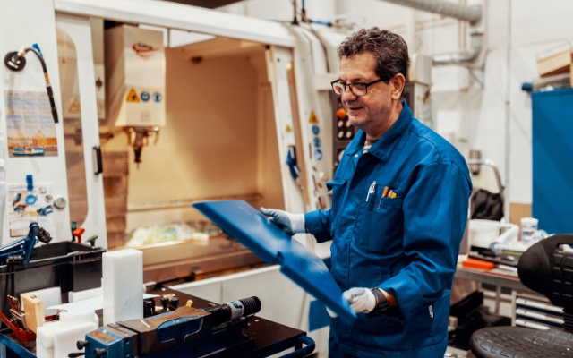 A smiling factory worker in a blue uniform and safety gloves holds two blue plastic parts inside a manufacturing workshop. Behind him is a CNC machine with an open door, and the workspace is organized with tools, posters, and components.
