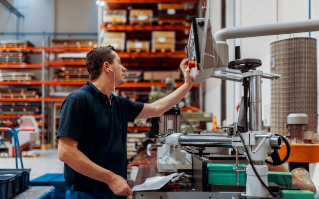 A man operating a machine in a warehouse or industrial workshop. He is wearing earphones and a dark polo shirt, and is pressing a button on a control panel. The background shows organized shelves filled with boxes and equipment.