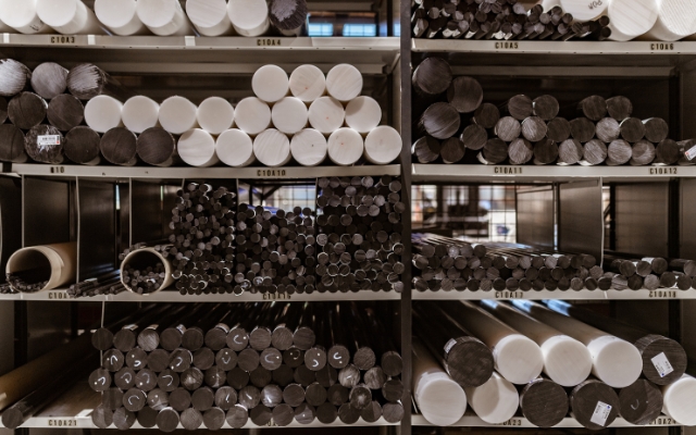 Förvaringshyllor fyllda med runda plaststavar i olika diametrar och färger, främst svarta och vita. Stavarna är prydligt organiserade i horisontella staplar, och varje hyllplan är märkt med en hyllkod. Miljön verkar vara ett industriellt lager.