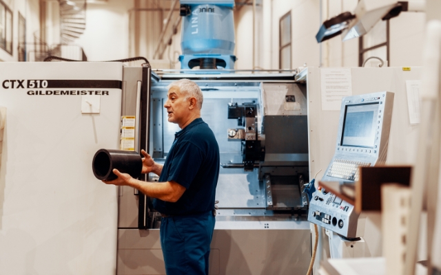 En man står framför en CNC-maskin märkt 'CTX 510 Gildemeister' och håller i ett cylindriskt svart arbetsstycke. Han verkar förklara eller inspektera delen. Den industriella maskinen bakom honom har en öppen kammare och ett kontrollpanel med skärm och tang