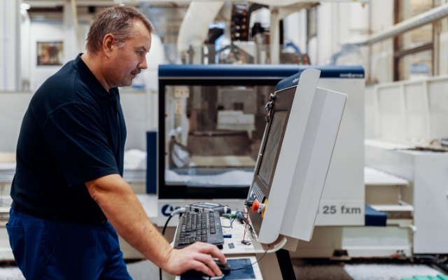 En man som arbetar vid ett datorstyrt gränssnitt framför en CNC-maskin i en industriell anläggning. Han är fokuserad på skärmen och använder tangentbord och mus för att styra utrustningen. I bakgrunden syns en stor automatiserad maskin märkt '25 fxm'.