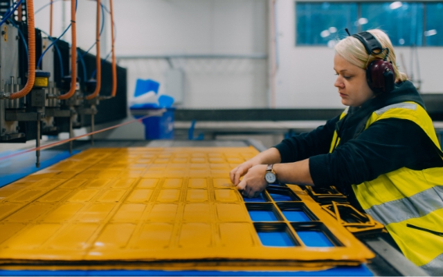 En kvinna i varselväst och hörselskydd arbetar vid en industriell maskin. Hon tar bort utskurna delar från en stor gul plastskiva som bearbetas av en CNC- eller laserskärmaskin. Miljön är en modern fabrik eller verkstad.