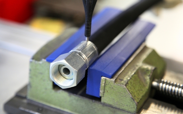Close-up of a metal hose fitting secured in a bench vise with blue protective jaws. A precision tool or probe is positioned over the fitting, likely for measurement or marking in a workshop or manufacturing setting.
