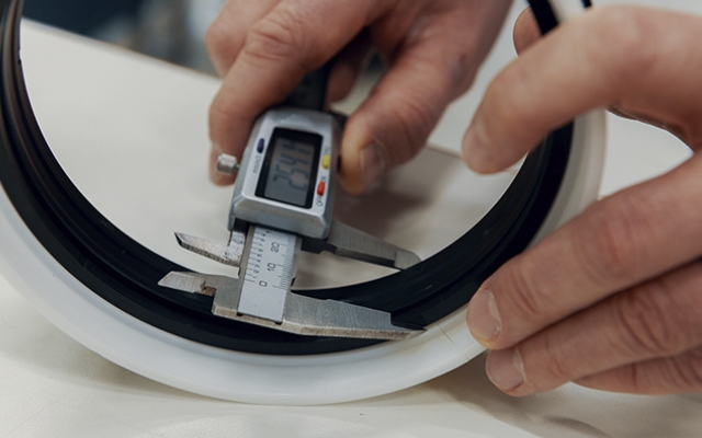 Close-up of hands using a digital caliper to measure the thickness of a black rubber or plastic ring seal. The caliper display shows a precise measurement, and the component is held steady on a white surface.