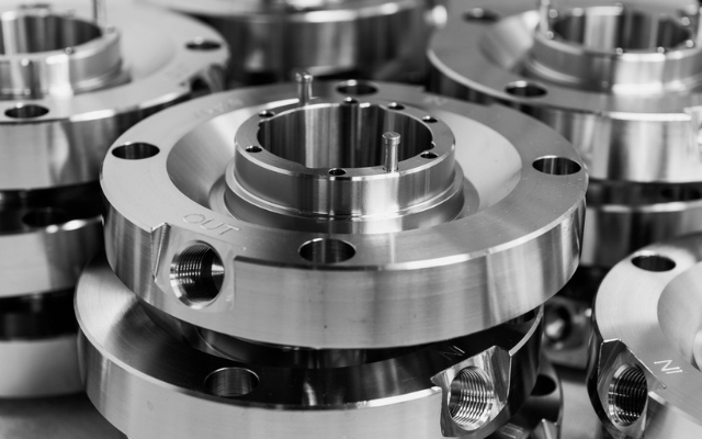 Close-up of precision-machined metal components with a polished finish. The parts feature threaded holes, circular flanges, and alignment pins, indicating use in industrial or mechanical assemblies. The image is in black and white.