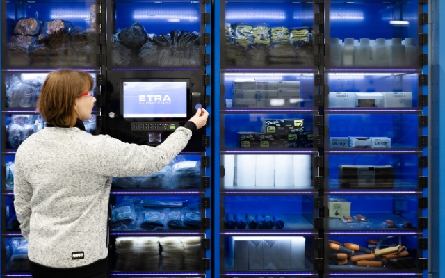 En kvinna står framför ett automatiserat lager- eller utdelningsskåp och använder en bricka eller nyckel för att öppna en lucka. Skåpet är fullt med arbetsutrustning som handskar, markörer och andra förnödenheter. 