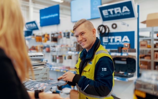 En leende ETRA-anställd i gul varselväst hjälper en kund vid en servicestation. Den anställde håller i en broschyr och i bakgrunden syns hyllor med verktyg och produkter samt flera ETRA-skyltar.