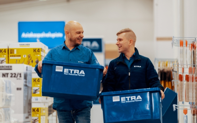 Två ETRA-anställda går leende bredvid varandra i en butik- eller lagerliknande miljö, båda bär varsin stor blå låda med ETRA-logotypen och texten ”Your Industrial Partner”. De verkar föra ett vänligt samtal.
