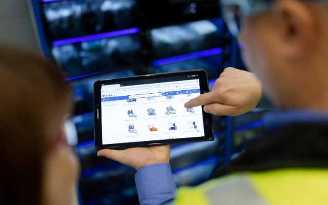 Two people are viewing a tablet displaying ETRA’s online store page with different types of work gloves. One person is pointing at the screen while shelves with products are visible in the background.