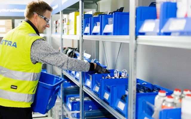 An ETRA employee wearing a high-visibility vest and safety glasses is picking items from blue storage bins on a shelf. He is holding a blue bin and appears to be organizing or collecting products in an industrial supply area.