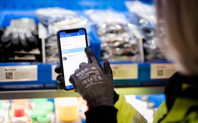 A person wearing work gloves is using a smartphone to place an order via ETRA’s digital interface. The screen shows product details for work gloves, and labeled bins are visible in the background.