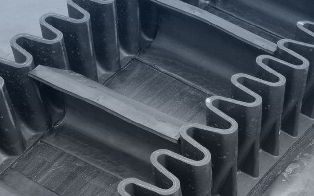 Close-up of a black corrugated rubber conveyor belt with sidewalls and cleats, designed for transporting bulk materials at steep angles.