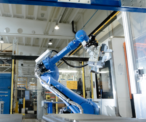 En bl&aring; industrirobot (Yaskawa) med robotarm arbetar i en produktionsanl&auml;ggning, omgiven av skyddsstaket och varningsskyltar.