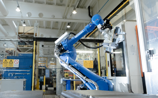 En bl&aring; industrirobot (Yaskawa) med robotarm arbetar i en produktionsanl&auml;ggning, omgiven av skyddsstaket och varningsskyltar.