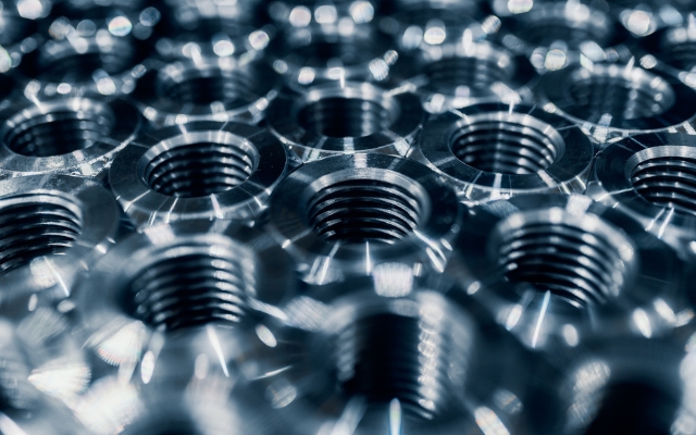 Close-up view of multiple shiny, machined metal nuts or threaded components arranged in a uniform pattern, reflecting light from their polished surfaces.
