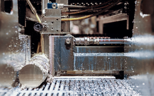 Närbild av en industriell metallkapmaskin i drift, som skär genom en cylindrisk metallstång med kylvätska och metallspån synliga på maskinen och arbetsytan.